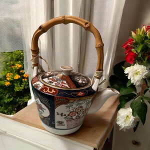 May include: A decorative ceramic teapot with a bamboo handle. The teapot features a detailed floral and geometric design in shades of red, blue, and gold. The handle is made of curved bamboo, adding a natural element to the design.