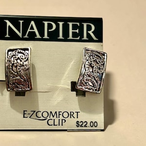 May include: Silver-toned Napier clip-on earrings with an ornate, scrolling design. The earrings are rectangular and feature a detailed, textured pattern. The card behind the earrings has the Napier logo and the text "E-Z COMFORT CLIP".