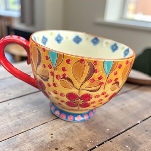 Pu&ograve; includere: Una grande tazza da t&egrave; decorativa in ceramica con manico e base rossi. La tazza presenta uno sfondo giallo con motivi floreali e a foglie in rosso, marrone e turchese. Il bordo &egrave; bordato da accenti floreali blu.