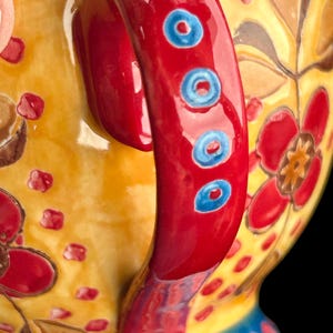 Pu&ograve; includere: Primo piano di una teiera in ceramica decorativa. Il manico &egrave; rosso brillante con dettagli circolari blu. La teiera presenta un corpo giallo con motivi floreali rossi. La base ha un design blu e giallo.