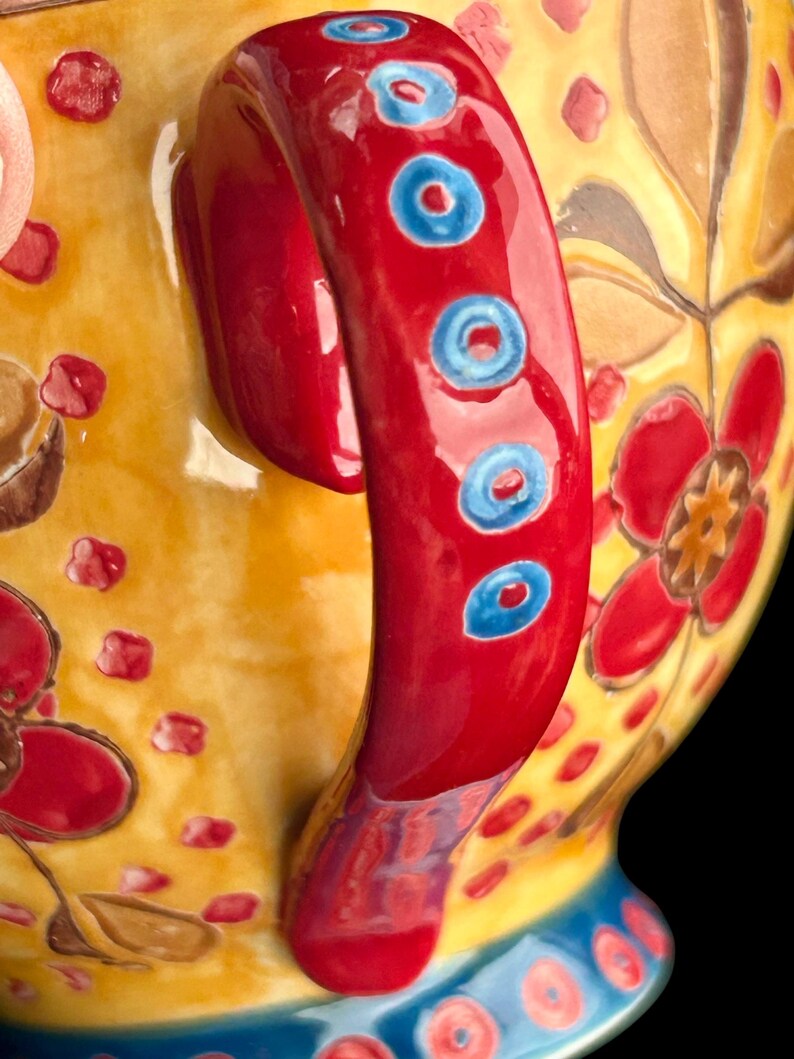 Pu&ograve; includere: Primo piano di una teiera in ceramica decorativa. Il manico &egrave; rosso brillante con dettagli circolari blu. La teiera presenta un corpo giallo con motivi floreali rossi. La base ha un design blu e giallo.
