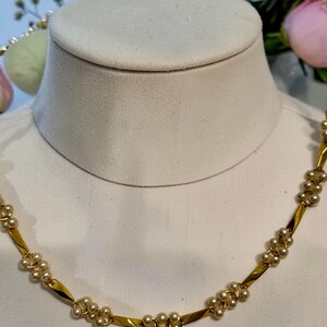 May include: A gold-toned necklace with small, clustered pearls. The necklace is displayed on a white mannequin bust. The design features alternating sections of gold and pearl clusters, creating a delicate and elegant look. The background includes flowers.
