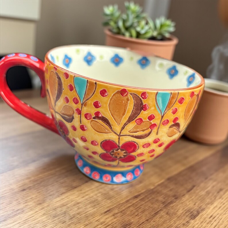Pu&ograve; includere: Una grande tazza in ceramica dipinta a mano con manico rosso e design floreale. La tazza presenta uno sfondo giallo con accenti marroni, rossi e blu. La tazza &egrave; appoggiata su una superficie di legno.