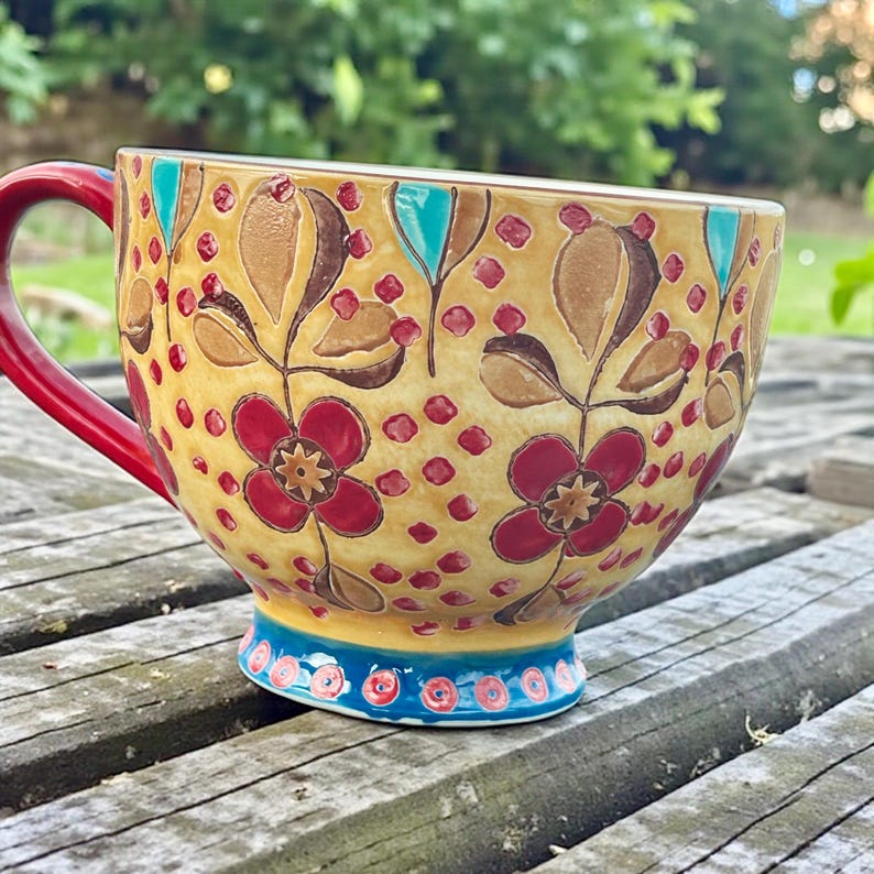 Pu&ograve; includere: Una grande tazza in ceramica con manico rosso e design floreale. La tazza presenta uno sfondo giallo con foglie marroni, fiori rossi e accenti turchese. La base ha un motivo blu e rosso.