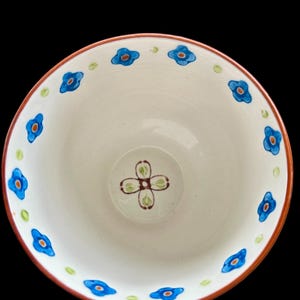 Pu&ograve; includere: Una ciotola in ceramica bianca con un motivo floreale. Il bordo &egrave; delineato in un colore marrone-rossastro. Fiori blu con centri arancioni sono distanziati attorno al bordo. Il centro della ciotola presenta un motivo floreale a quattro petali in marrone e verde.