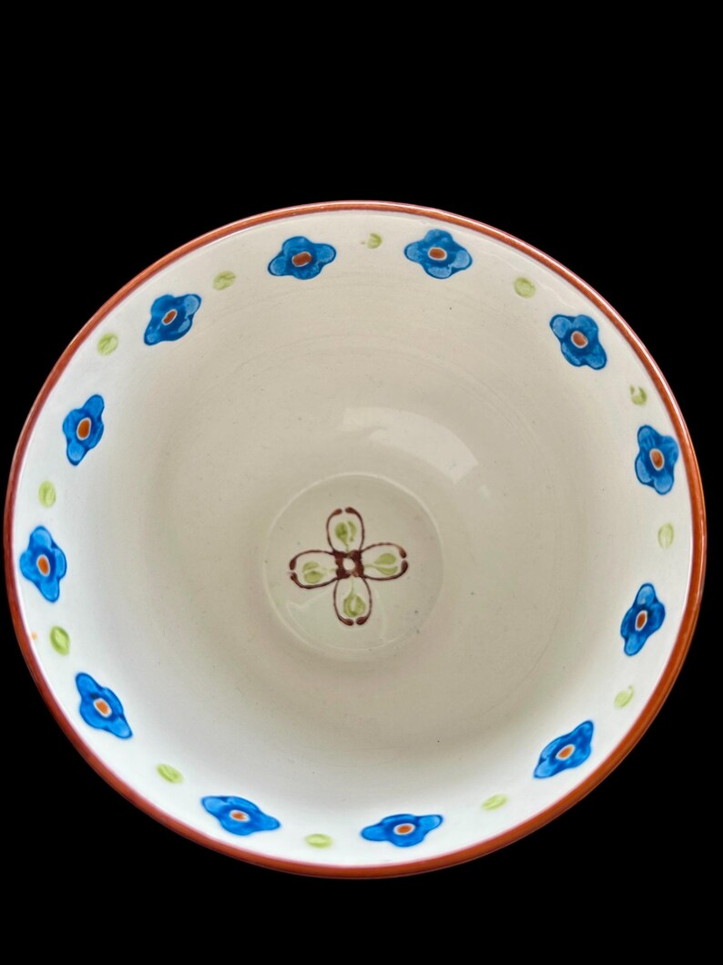 Pu&ograve; includere: Una ciotola in ceramica bianca con un motivo floreale. Il bordo &egrave; delineato in un colore marrone-rossastro. Fiori blu con centri arancioni sono distanziati attorno al bordo. Il centro della ciotola presenta un motivo floreale a quattro petali in marrone e verde.