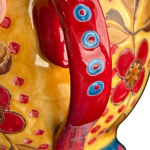 Pu&ograve; includere: Primo piano di una tazza in ceramica con un vivace motivo floreale. Il manico &egrave; rosso lucido con accenti blu. La tazza presenta uno sfondo giallo con motivi floreali rossi e marroni e una base blu.