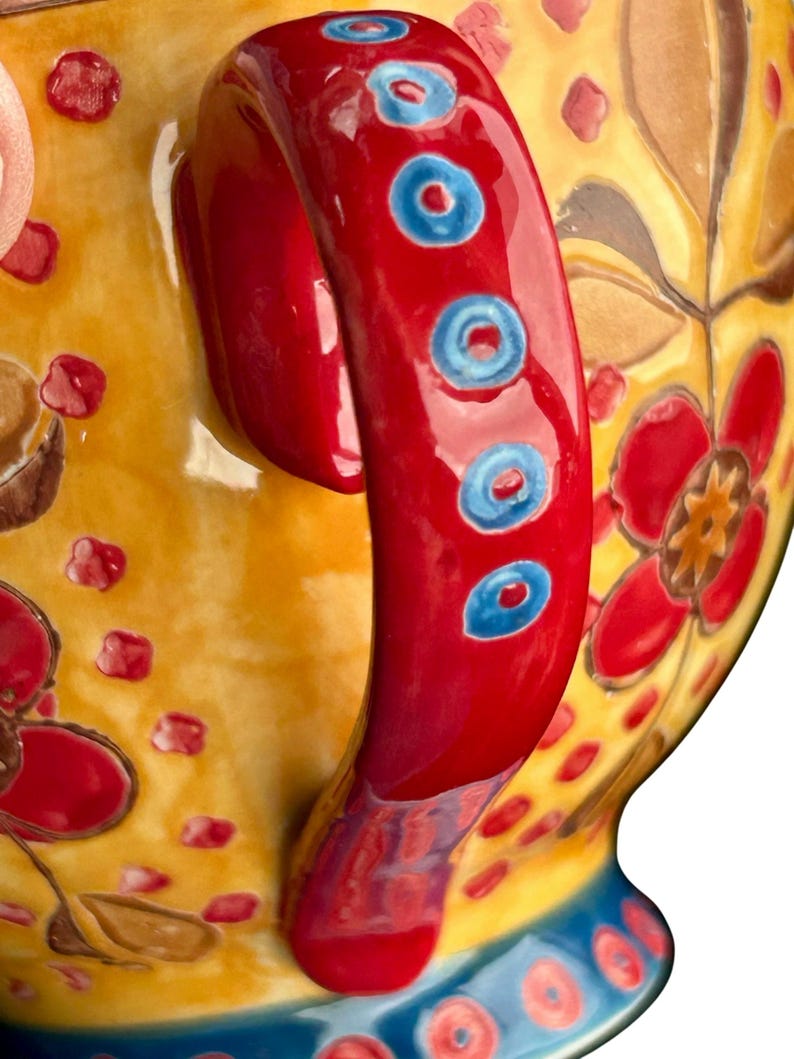 Pu&ograve; includere: Primo piano di una tazza in ceramica con un vivace motivo floreale. Il manico &egrave; rosso lucido con accenti blu. La tazza presenta uno sfondo giallo con motivi floreali rossi e marroni e una base blu.