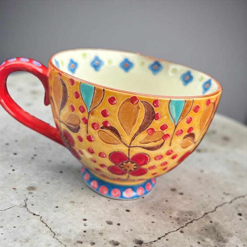 Pu&ograve; includere: Una tazza da t&egrave; in ceramica dipinta a mano con manico e base rossi. La tazza presenta uno sfondo giallo con disegni floreali e di foglie in rosso, marrone e turchese. Il bordo e la base hanno accenti blu.