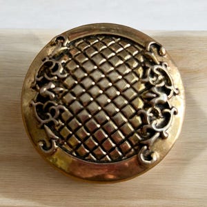 May include: A round, gold-toned metal button with a decorative, embossed design. The button has a raised, geometric pattern in the center and a floral design around the edge.