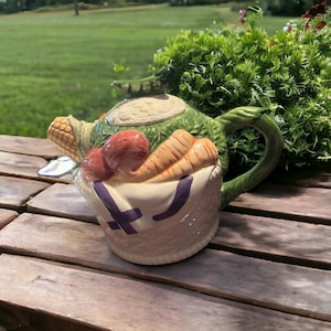May include: A ceramic teapot shaped like a basket filled with vegetables. The teapot is decorated with a green pea pod, corn, carrots, and red tomatoes. The teapot has a white base with purple accents.
