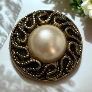 May include: A gold-toned button with a large, white pearl in the center. The button is decorated with a swirling, intricate design.