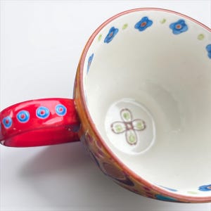 Pu&ograve; includere: Una tazza in ceramica con manico rosso e interno bianco. Il bordo &egrave; decorato con motivi floreali blu. Il manico presenta motivi circolari blu e rossi. L'esterno ha un design colorato e fantasia.