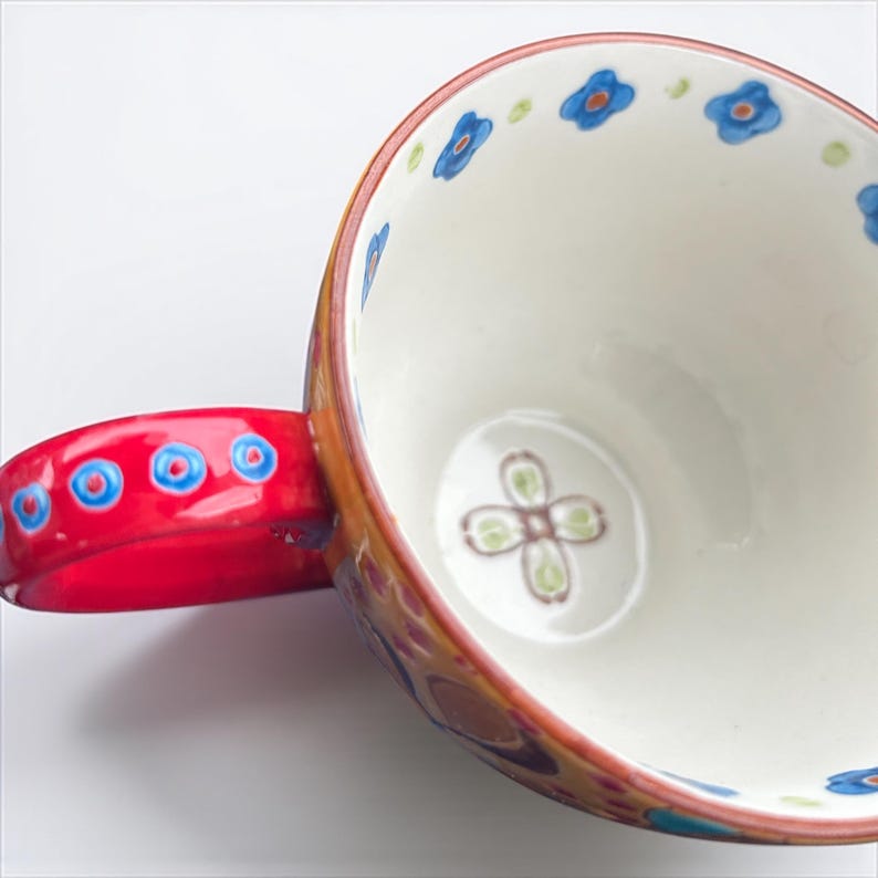Pu&ograve; includere: Una tazza in ceramica con manico rosso e interno bianco. Il bordo &egrave; decorato con motivi floreali blu. Il manico presenta motivi circolari blu e rossi. L'esterno ha un design colorato e fantasia.