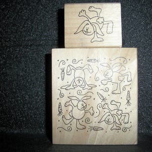 May include: Four wooden stamps with black line drawings of bunnies and carrots. The bunnies are in various poses, including jumping, standing, and sitting. The stamps are on a dark gray background.