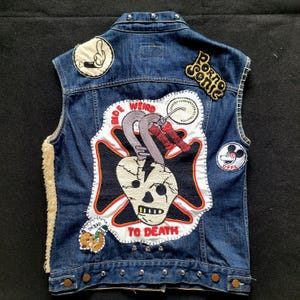 Peut inclure: Veste en jean sans manches ornée de divers écussons. Les écussons incluent une tête de mort, l'inscription "Ride Weird To Death" et des personnages de dessins animés. La veste a une doublure en peau de mouton et des boutons en métal.