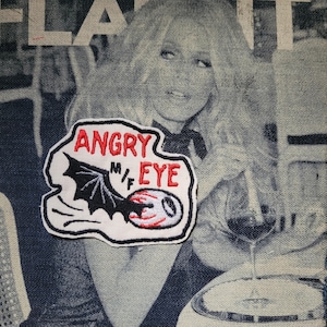 Peut inclure: Écusson brodé avec les mots "ANGRY M/F EYE" en lettres rouges. L'écusson présente une aile de chauve-souris noire et un motif d'œil rouge et blanc. L'écusson est sur un fond en denim avec une image en noir et blanc d'une femme.