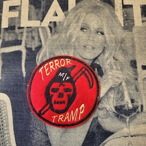 Peut inclure: Un écusson rond rouge avec les mots "TERROR TRAMP" en lettres dorées. L'écusson présente un motif de crâne de la Faucheuse noir avec une faux. Le fond est un tissu en jean avec une image en noir et blanc.