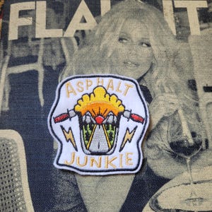 Op de afbeelding: Geborduurde witte patch met de woorden "ASPHALT JUNKIE" en een motorfietsontwerp. Het ontwerp bevat een gele zon, een weg en bliksemschichten. De patch staat op een denim achtergrond.