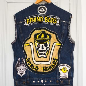 Peut inclure: Un gilet en jean bleu foncé orné de plusieurs écussons. Le dos présente un grand écusson avec un motif de crâne et les mots "WORLD NOMADS" et "BEHIND BARS". D'autres écussons incluent un logo Harley Davidson et un personnage de dessin animé jaune avec les mots "FUCK YOU HIPPIES".
