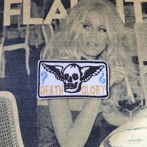 Puede incluir: Parche rectangular con un diseño de calavera y alas, las palabras "DEATH GLORY" en letras doradas y dos signos de interrogación azules. El parche es blanco con un borde negro y está sobre un fondo de mezclilla.