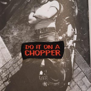 Peut inclure: Un écusson rectangulaire noir brodé avec le texte rouge DO IT ON A CHOPPER. L'écusson présente une bordure noire et est placé sur une photographie en noir et blanc montrant une personne assise sur une moto avec le texte visible.