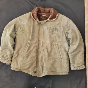 Puede incluir: Una chaqueta de trabajo verde forrada con cuello de piel sintética marrón. La chaqueta tiene un parche en el pecho izquierdo con las letras "MN" y un ancla.