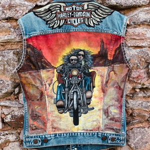 Peut inclure: Un gilet en jean avec une scène peinte de deux personnes conduisant une moto à travers un paysage désertique. Le gilet a un logo Harley-Davidson avec des ailes au dos.