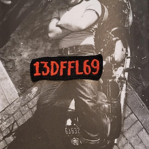 Peut inclure: Un écusson rectangulaire brodé noir avec le texte 13DFFL69 en fil rouge vif. L'écusson est placé sur une photographie en noir et blanc montrant une personne portant une casquette et un t-shirt, assise sur une moto avec une plaque d'immatriculation.