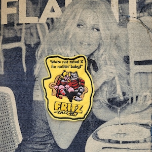 Op de afbeelding: Een gele patch met de stripfiguren Fritz the Cat en een kat op een bruine bank. De patch heeft de tekst "We're not rated X for nothin' baby!" en "FRITZ the CAT" in het geel.