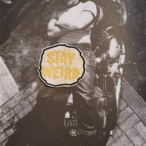 Op de afbeelding: Een ronde geborduurde patch met de gele tekst STAY WEIRD en een dikke zwarte rand. De patch is geplaatst over een zwart-witfoto van een persoon op een motorfiets, gekleed in een pet en t-shirt, met de tekst 63632 duidelijk zichtbaar.