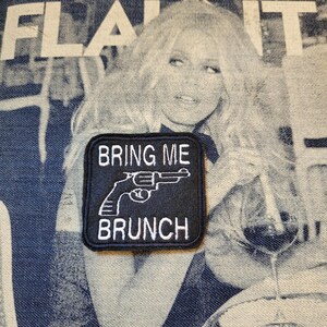 Może przedstawiać: Czarna kwadratowa naszywka z białym haftowanym tekstem i grafiką rewolweru. Tekst brzmi "BRING ME BRUNCH". Naszywka jest umieszczona na dżinsowym tle z wyblakłym wizerunkiem kobiety.