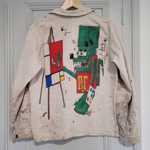 May include: A white denim jacket with a colorful painting of a cartoon monster holding a paintbrush and painting a canvas. The monster is green, red, and yellow. The canvas is blue, red, yellow, and white. The jacket has paint splatters on it.