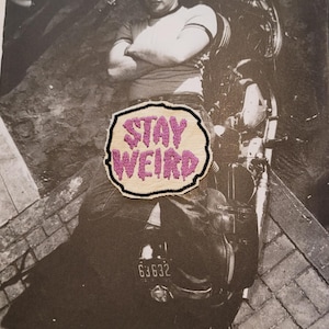 Könnte beinhalten: Ein gestickter Aufnäher mit cremefarbenem Hintergrund und schwarzem Rand, der den Text STAY WEIRD in strukturiertem, leuchtend pinkfarbenem Garn zeigt. Der Aufnäher liegt auf einem Schwarz-Weiß-Foto einer Person auf einem Motorrad.