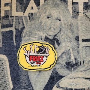 May include: A yellow patch featuring a black cat in a bowl, with the red text "Fritz the Cat" and legs. The patch is set against a denim background with a monochrome image of a woman.