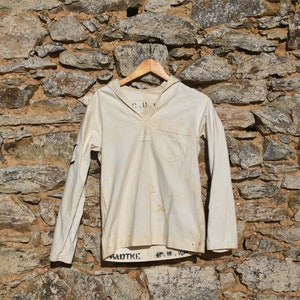 May include: A white long-sleeved shirt with a sailor collar, hanging on a wooden hanger against a stone wall. The shirt has a pocket on the left side and the text "C.D.V." and "RADTKE" printed on it.