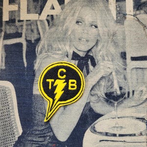 Puede incluir: Un parche bordado en negro y amarillo con las letras "C T B" y un diseño de rayo. El parche tiene forma de bocadillo. El fondo es una tela tipo denim con una imagen en blanco y negro de una mujer.