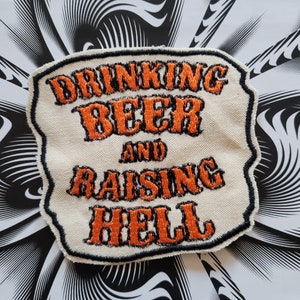 Peut inclure: Un patch en tissu blanc avec du texte brodé en noir et orange qui dit "Drinking Beer and Raising Hell".