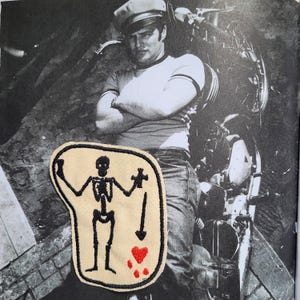 Op de afbeelding: Een beige geborduurde patch met een zwart skeletfiguur dat een klein kruis vasthoudt, met een naar beneden wijzende pijl en een rood hartje eronder, alles omlijnd met zwart garen, geplaatst over een zwart-witfoto van een persoon.