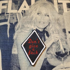 Peut inclure: Écusson noir en forme de losange avec bordure blanche et texte rouge brodé "Don't give a fuck". L'écusson est posé sur un fond en denim avec une image estompée d'une femme.