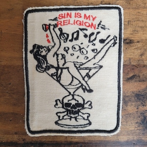 Könnte beinhalten: Ein weißer gestickter Aufnäher mit schwarzen Nähten. Der Aufnäher zeigt eine Frau in einem Martini-Glas mit einem Totenkopf und gekreuzten Knochen am Boden. Der Text "SIN IS MY RELIGION" ist in Rot über dem Bild geschrieben.