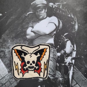 Puede incluir: Parche bordado con una calavera y huesos cruzados, alas de murciélago negras y detalles rojos y naranjas. Rayos dorados flanquean el diseño de la calavera. El parche está sobre un fondo crema.