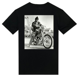 Puede incluir: Foto en blanco y negro de un hombre con una camisa a rayas y un sombrero montando una motocicleta. La motocicleta tiene un tubo de escape largo y recto.