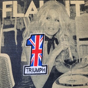 Peut inclure: Tissu en jean avec une image en noir et blanc d'une femme et le mot "FLAUNT" en blanc. Un écusson avec le drapeau Union Jack, le chiffre "1" et le mot "TRIUMPH" est cousu sur le tissu.