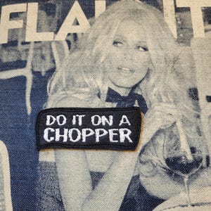 Peut inclure: Un écusson noir brodé avec le texte "DO IT ON A CHOPPER" en lettres blanches. L'écusson est posé sur un fond en jean avec une image estompée d'une femme et le mot "FLAUNT" en blanc.