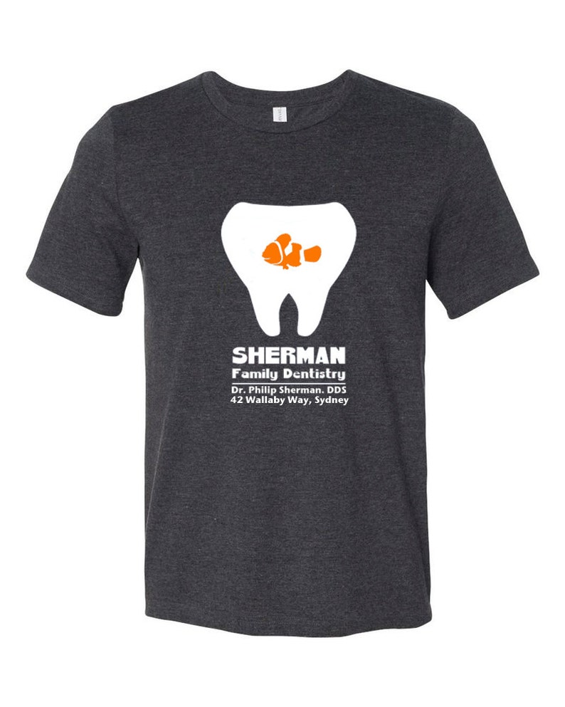 Puede incluir: Una camiseta gris oscuro con un gr&aacute;fico de diente blanco y un pez payaso naranja y blanco dentro del diente. El texto "SHERMAN Family Dentistry Dr. Phillip Sherman, DDS 42 Wallaby Way, Sydney" est&aacute; impreso debajo del gr&aacute;fico.