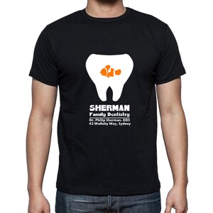Puede incluir: Camiseta negra con un gr&aacute;fico blanco de un diente con un pez de dibujos animados naranja y blanco en su interior. El texto "SHERMAN Family Dentistry Dr. Phillip Sherman, DDS 42 Wallaby Way, Sydney" est&aacute; impreso en blanco debajo del gr&aacute;fico.