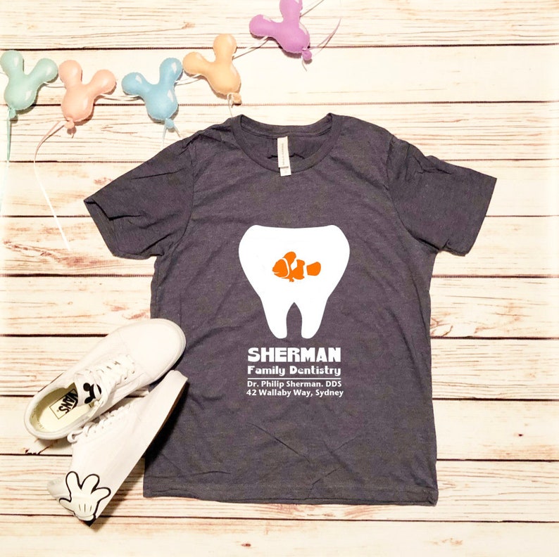 Puede incluir: Una camiseta gris oscuro con un gr&aacute;fico de diente blanco y un pez payaso naranja y blanco dentro del diente. El texto "SHERMAN Family Dentistry Dr. Phillip Sherman, DDS 42 Wallaby Way, Sydney" est&aacute; impreso debajo del diente.