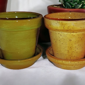 May include: Two ceramic flower pots with matching saucers. One pot is olive green, the other is speckled yellow. Both are empty. Additional red and green pots are in the background. Succulents are on either side.