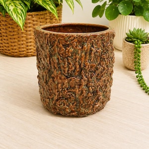 May include: A brown ceramic planter with a textured, bark-like exterior. The cylindrical pot has a wide opening and is suitable for indoor or outdoor plants. The planter is displayed on a light wood surface, with other plants in the background.